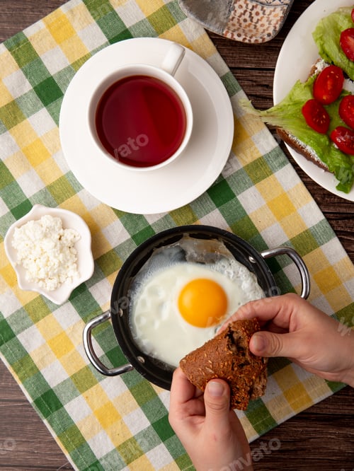 Preview: top view of woman hands holding slice of bread with fried egg, tea, cottage cheese on cloth surface