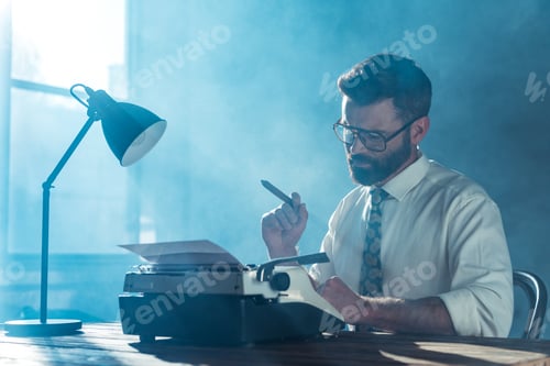 Preview: bearded journalist in glasses sitting at table, holding cigar and looking at vintage typewriter near