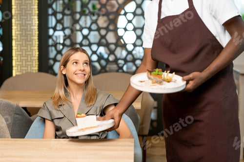 Preview: Young African water in uniform placing plate with dessert on table by blond girl