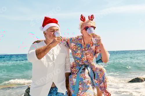 Preview: Funny couple wearing christmas hats drinking sparkling wine and enjoying christmas on tropical beach