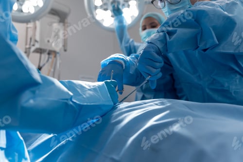 Preview: Diverse group of male and female surgeons in operating theatre wearing face masks performing surgery