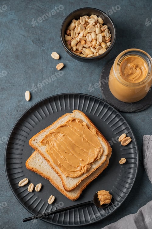 Preview: Peanut butter toast on a black plate on a dark background with nuts, napkin and spoon.