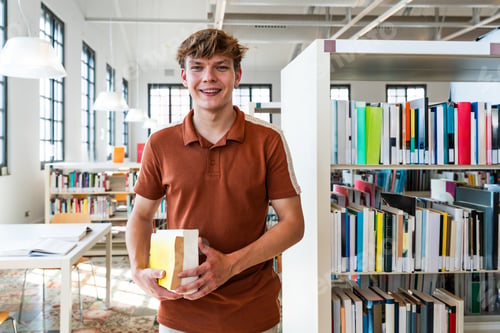 Preview: Smiling student holding books standing in library