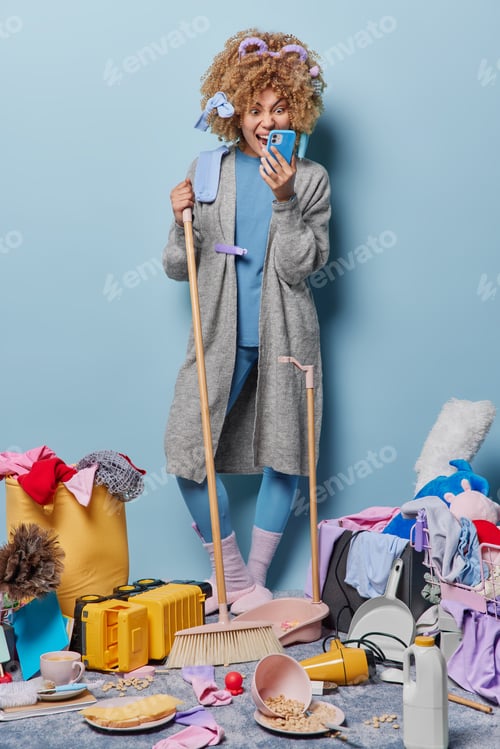 Preview: Crazy curly haired housewife stares in smartphone screen reacts to received message cleans house