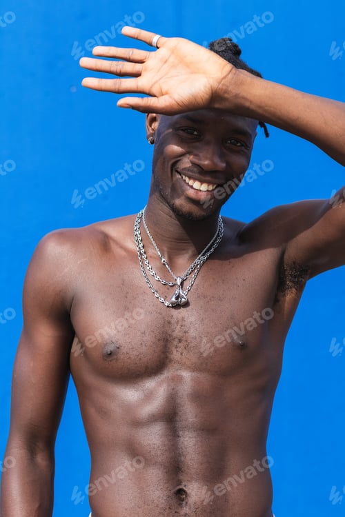 Preview: Black man standing shirtless over isolated blue background smiling to the camera and covering his