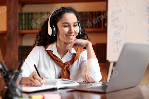 Preview: Woman sitting at table, using laptop and writing in notebook