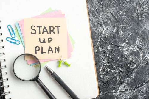 Preview: top view start up plan note on sticker with pen and magnifier on gray background lesson student