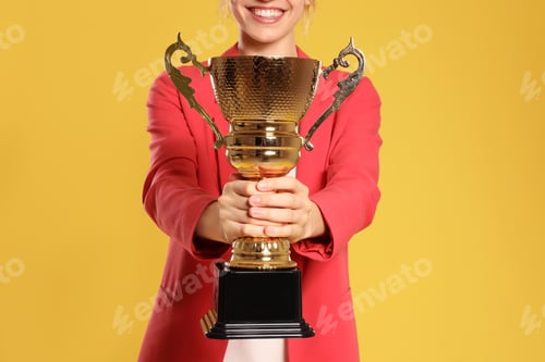 Preview: Young businesswoman with gold trophy cup on yellow background, closeup