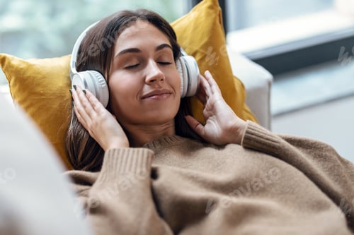 Preview: Beautiful woman listening to music with headphone while using smartphone lying on sofa at home.