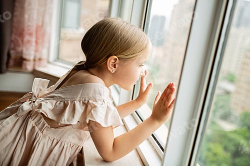 Preview: Beautiful Caucasian girl with blonde hair looking through the window at home during quarantine