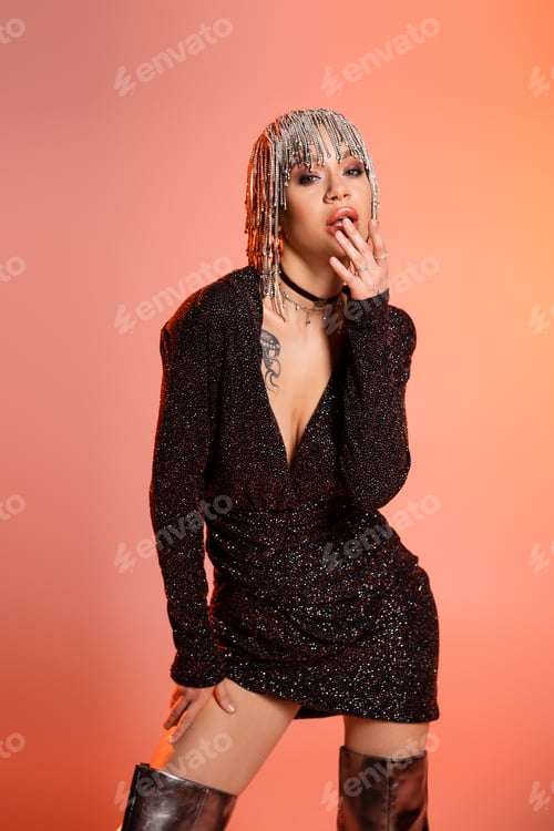 Preview: sexy woman in black dress and metallic headwear touching lips and looking at camera on coral pink