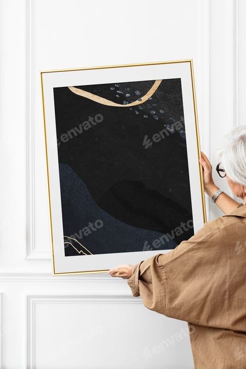 Preview: Senior woman hanging an aesthetic picture frame on a white wall