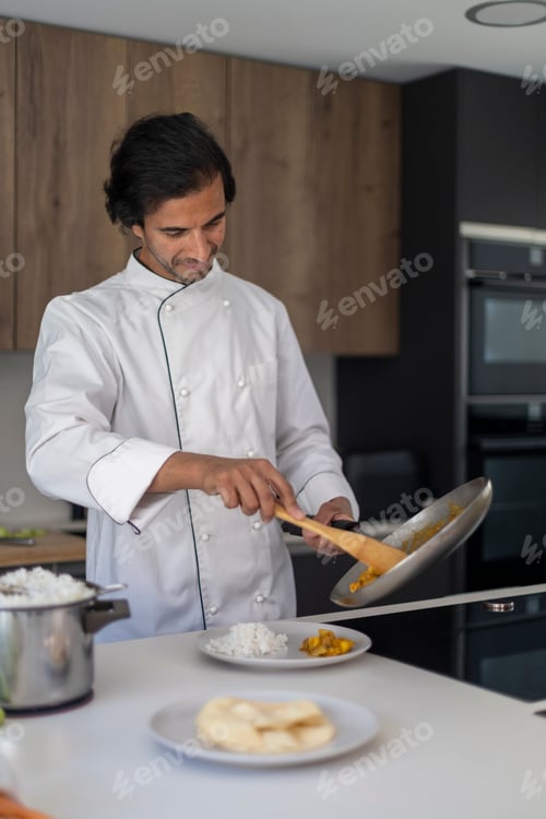 Preview: Chef in white uniform serving food from a pan onto a plate in a modern kitchen.