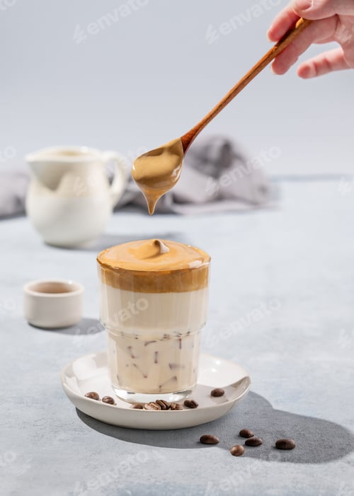 Preview: Dalgona coffee. A hand holds a spoon with whipped foam, for instant coffee with ice and milk