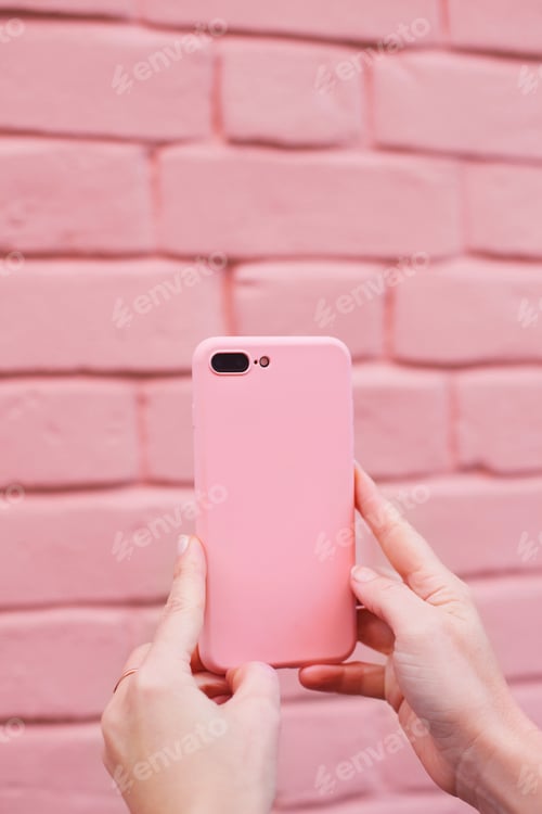 Preview: Woman holding pink mobile phone on a pink brick wall background close up
