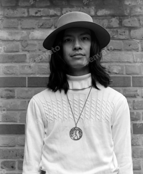 Preview: Young asian man wearing hat and pendant posing in front of brick wall in monochrome analog photo