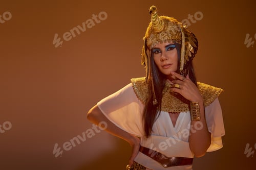 Preview: Stylish woman in Egyptian attire and headdress touching chin and posing on brown background