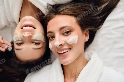 Preview: Two women in bathrobes lying near in bed with cosmetic mask on faces happily looking in camera