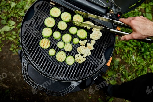 Preview: Hand of young man grilling zucchini vegetable on huge gas grill .
