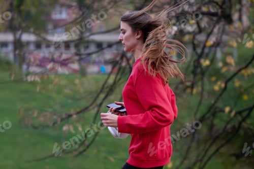 Preview: Young beautiful woman running in autumn park and listening to music with headphones on smartphone