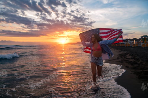 Preview: Beautiful patriotic young woman with the American flag held in her standing in the summer sunset