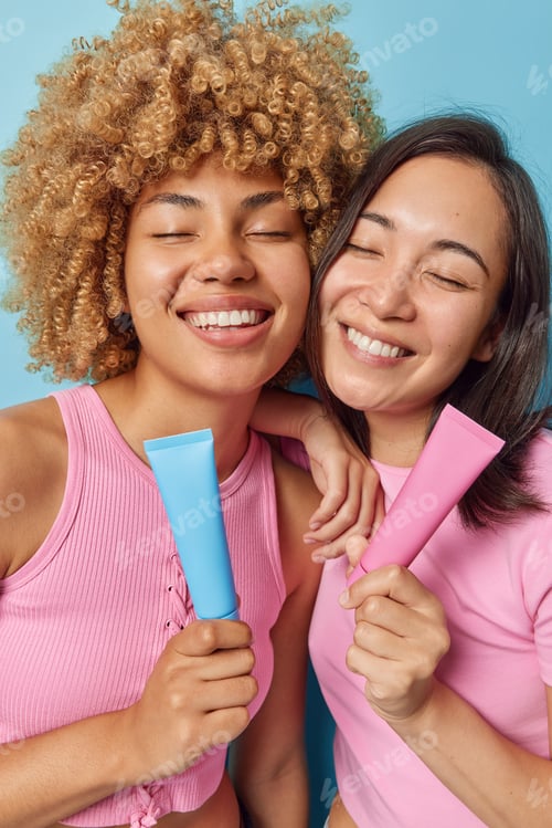 Preview: Vertical shot of cheerful mixed race young women keep eyes closed smile broadly hold toothpaste tubs