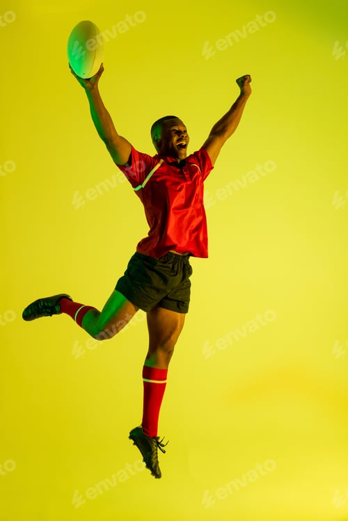 Preview: Happy african american male rugby player raising hands over yellow lighting
