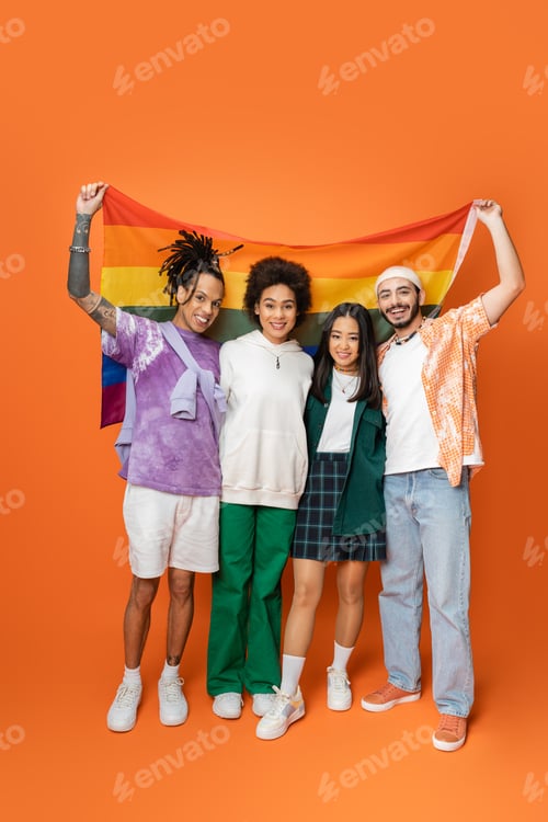 Preview: full length of smiling multiethnic friends in trendy clothes holding lgbt flag on orange