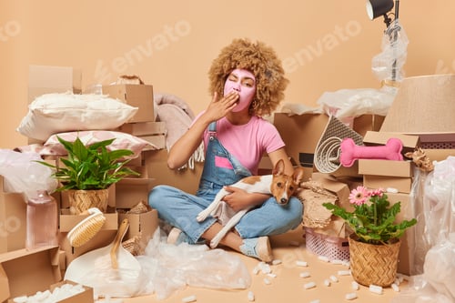Preview: Tired sleepy curly haired woman covers mouth as yawns poses in messy room with dog undergoes beauty