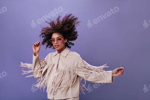 Preview: Curly woman in glasses posing on purple background. Pretty young curly girl in beige jacket dancing