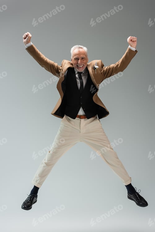 Preview: full length of happy middle aged groom in suit with floral boutonniere jumping on grey