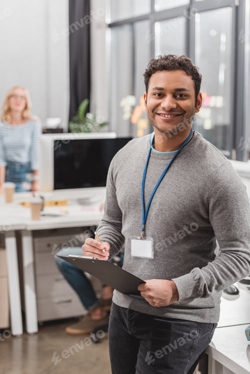 Preview: happy african american man with name tag writing something on planchette at modern office
