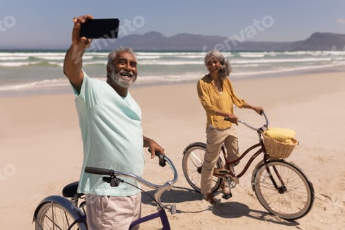 Preview: Front view of happy senior couple talking selfie with mobile phone on beach in the sunshine