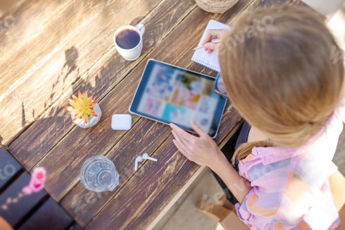 Preview: Overhead woman whiting notes and looking on digital tablet, dark wooden table, coffee and water