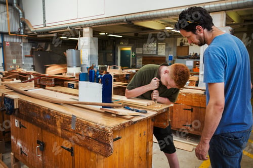 Preview: Two men standing at a workbench in a boat-builder's workshop, working on a wooden joint.