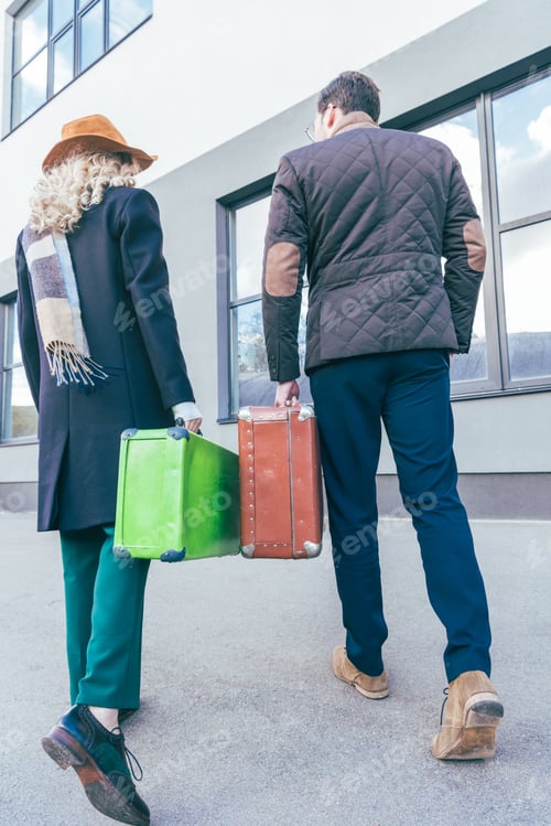 Preview: back view of stylish young couple holding suitcases and walking on street
