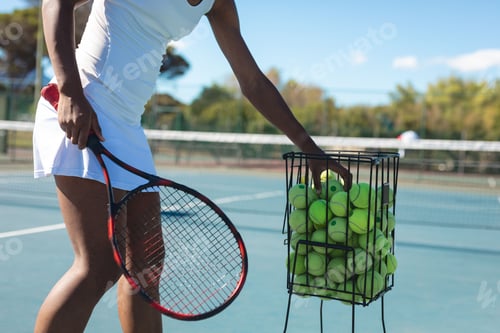 Preview: African American woman tennis player picking balls from basket on blue court, copy space