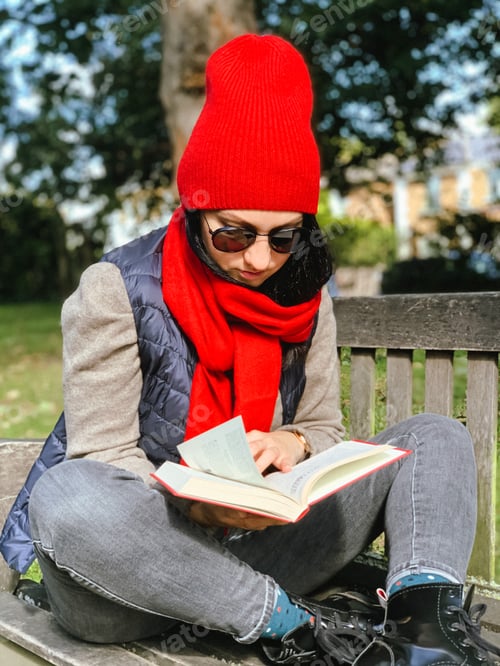 Preview: Young candid woman in casual autumnal clothes siting on a wooden bench in park and reading red book