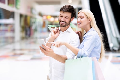 Smiling Couple Using Cellphone Searching Sales Offers Standing In Mall
