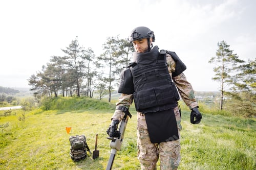 Preview: A man in a military uniform and bulletproof vest works in the forest with a metal detector