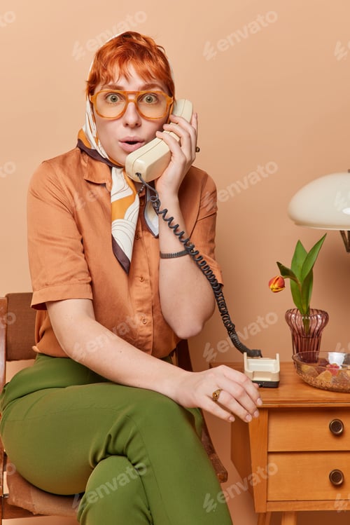Preview: Redhead shocked redhead woman wears big spectacles kerchief shirt and trousers talks on old phone