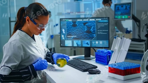 Preview: Chemist with protection glass examining various bacteria holding petri dish