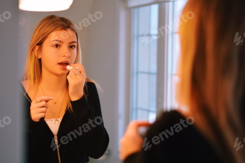 Preview: Young teenage girl applying lipstick, lip balm in front of a bathroom mirror
