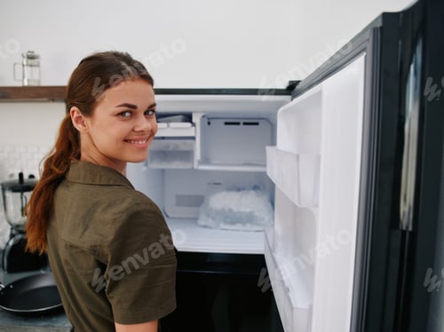 Preview: Woman smiling with teeth looking into camera in kitchen at home opened freezer empty with ice inside