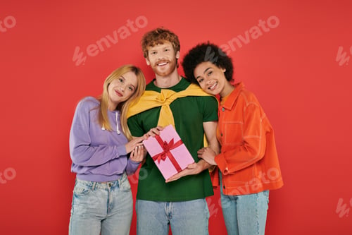 Preview: open relationship, polygamy, cheerful redhead man holding gift box near interracial bisexual women,