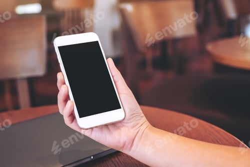 Vista previa: A hand holding mobile phone with blank desktop screen with laptop computer on the table