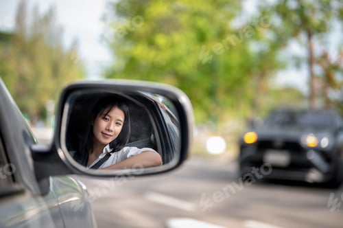 Preview: Side mirror shot of smiling asian woman in white shirt, driver of a car parked at lot or roadside.
