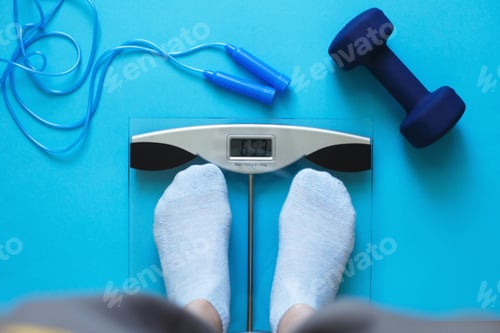 Preview: Woman measures her weight on scale on a blue background with skipping rope and dumbbells