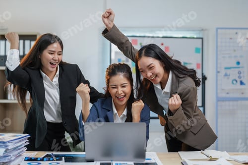 Preview: Three young businesswomen are raising their arms and smiling while looking at a laptop in an office,