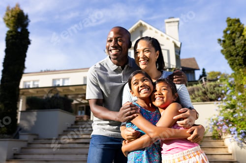 Preview: Outdoors, happy biracial family of four is smiling and hugging outside their home